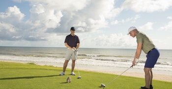 Two golfers standing on a green golf course directly next to a sandy beach and ocean waves in Northeast Florida.